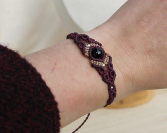 Puede incluir: Un brazalete de macramé de color morado oscuro y beige con una piedra preciosa negra en el centro.