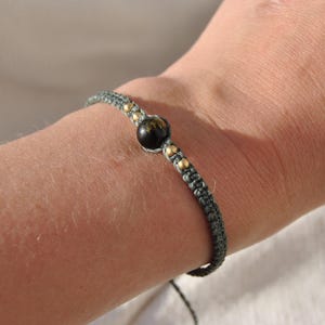 May include: A macrame bracelet with a dark grey cord. It features a central black bead and small gold-coloured beads. The bracelet is worn on a wrist.