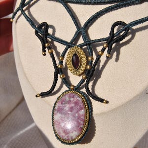 May include: A handcrafted macrame necklace featuring a large oval pink gemstone pendant with a gold-tone bezel. The necklace has a dark teal cord with gold-colored beads and a smaller oval pendant with a dark red stone.