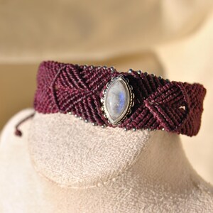 May include: A burgundy macrame bracelet with a central oval-shaped moonstone. The bracelet features a woven design with silver-colored beads along the edges. The moonstone has a silver bezel setting.