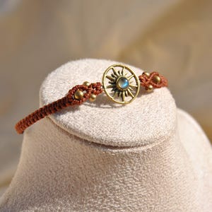 Pode incluir: Uma pulseira de macramé castanho com um berloque dourado em forma de sol com uma pedra azul central. A pulseira tem contas douradas e é exibida em uma superfície de tom neutro. Um acessório de estilo boêmio.