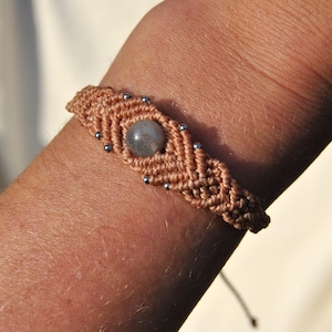 May include: A handmade macrame bracelet in a light brown color, featuring a central gray gemstone and small silver beads. The woven design is visible, showcasing the craftsmanship of the jewelry.