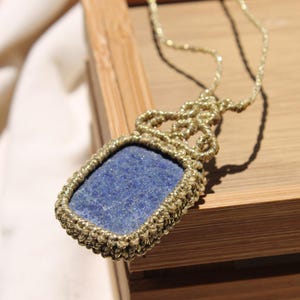 May include: A handmade necklace featuring a rectangular, deep blue stone pendant. The stone is encased in a woven, gold-colored macrame setting and hangs from a matching cord. The necklace rests on a wooden surface.