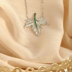 May include: A silver-toned necklace featuring a textured maple leaf pendant. The center vein of the leaf is embellished with a vertical row of small, vibrant green stones. The pendant hangs from a delicate, thin silver-toned metal chain.