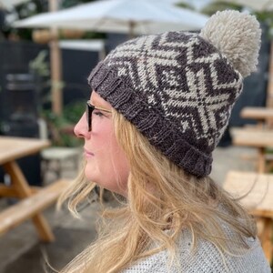 May include: A dark gray and cream-colored knit hat with a geometric pattern and a cream-colored pom-pom. The hat is worn by a person with blonde hair and glasses. The hat is a winter accessory.
