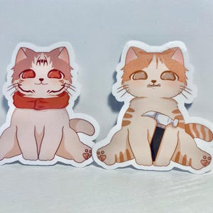 May include: Two cartoon cat stickers. One cat is wearing a red scarf and the other cat is holding a hammer.