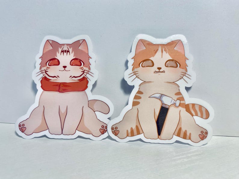 May include: Two cartoon cat stickers. One cat is wearing a red scarf and the other cat is holding a hammer.