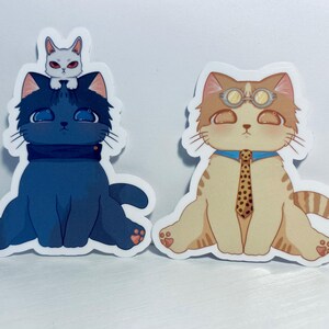 May include: Two cat stickers. One is a blue cat with a white cat on its head. The other is a tan cat wearing goggles and a tie.