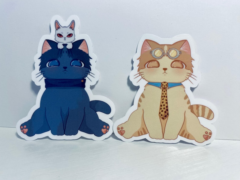 May include: Two cat stickers. One is a blue cat with a white cat on its head. The other is a tan cat wearing goggles and a tie.