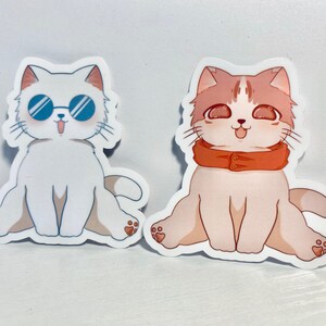 May include: Two cartoon cat stickers. One cat is white with blue sunglasses and the other is light pink with an orange scarf.