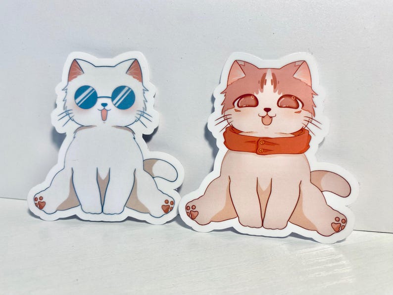 May include: Two cartoon cat stickers. One cat is white with blue sunglasses and the other is light pink with an orange scarf.