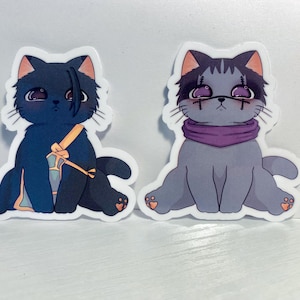May include: Two black and white cat stickers with cartoon faces. The cat on the left is wearing a purple and yellow scarf. The cat on the right is wearing a purple scarf.