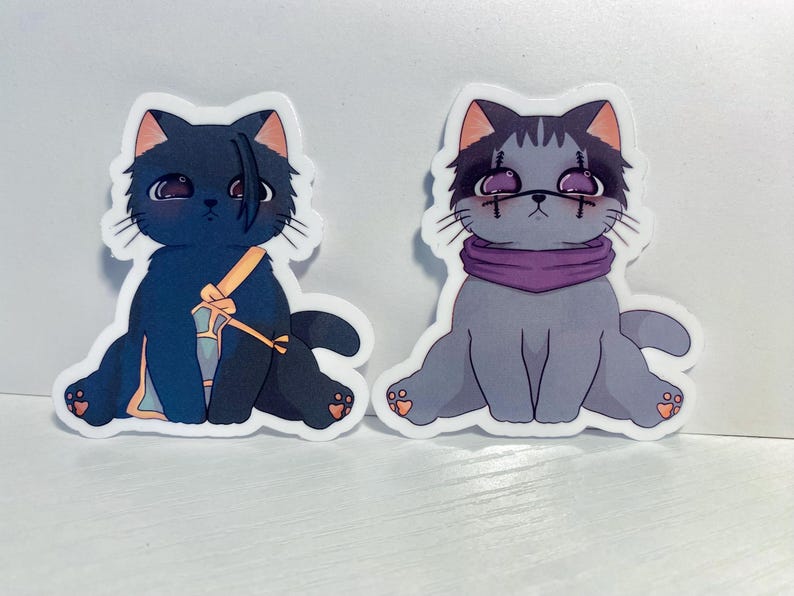 May include: Two black and white cat stickers with cartoon faces. The cat on the left is wearing a purple and yellow scarf. The cat on the right is wearing a purple scarf.