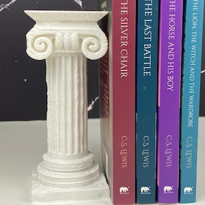 Puede incluir: Un sujetalibros blanco con forma de columna de estilo clásico se encuentra junto a una pila de libros. Los libros tienen lomos coloridos con títulos como "La silla de plata" y "El león, la bruja y el armario" de C.S. Lewis.