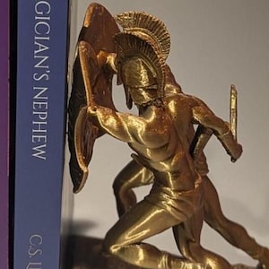 May include: Gold-colored bookends depicting two Spartan warriors in battle. One warrior holds a shield and the other holds a sword. The bookends are on a brown base.