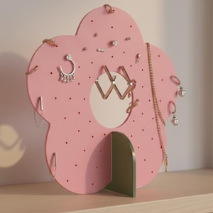 Puede incluir: Un organizador de joyas rosa en forma de flor con un espejo circular en el centro. Tiene pequeños agujeros para pendientes y ganchos para collares. La base es verde y se exhibe en una estantería de madera.