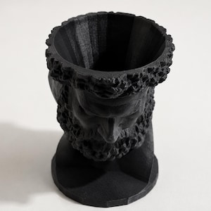 May include: A matte black planter shaped like a human head with a textured beard and hair. The top of the head is open, creating a cylindrical cavity for holding items. The sculpture features a circular base and a detailed facial structure.