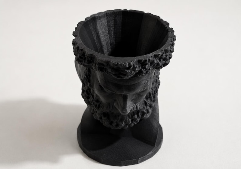 May include: A matte black planter shaped like a human head with a textured beard and hair. The top of the head is open, creating a cylindrical cavity for holding items. The sculpture features a circular base and a detailed facial structure.