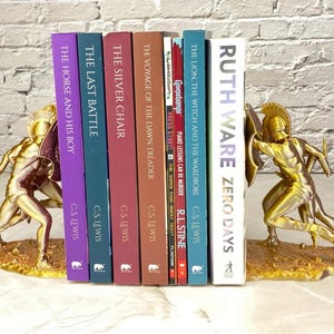 May include: Gold-colored bookends featuring a warrior design, holding a collection of books with titles like "The Horse and His Boy" and "Zero Days." The bookends are on a light-colored surface.