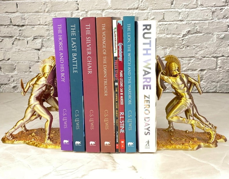 May include: Gold-colored bookends featuring a warrior design, holding a collection of books with titles like "The Horse and His Boy" and "Zero Days." The bookends are on a light-colored surface.