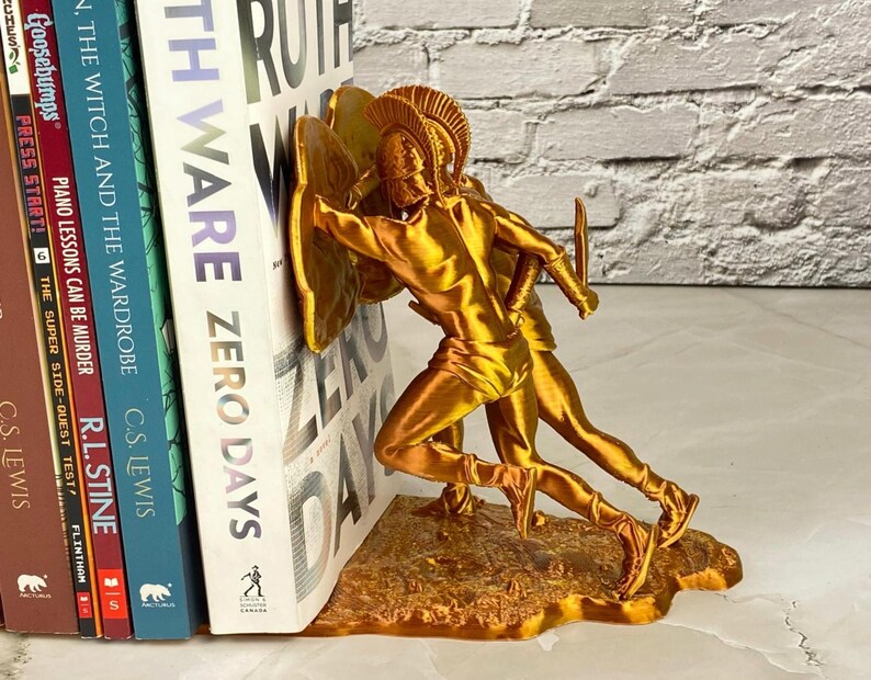 May include: A gold-colored bookend featuring two figures in classical armor, holding shields and swords. The bookend is designed to hold books upright. The books include titles like "The Witch and the Wardrobe" and "Zero Days".