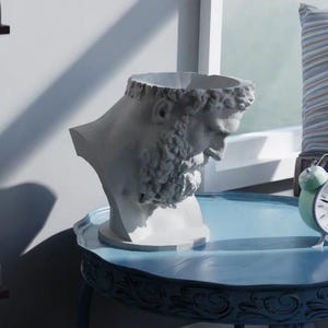 May include: A white bust planter shaped like a bearded man with curly hair and a downward gaze. The sculpture features a hollow top opening for plants and is positioned on a blue table next to a small, round, light green twin-bell alarm clock.