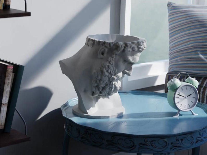 May include: A white bust planter shaped like a bearded man with curly hair and a downward gaze. The sculpture features a hollow top opening for plants and is positioned on a blue table next to a small, round, light green twin-bell alarm clock.