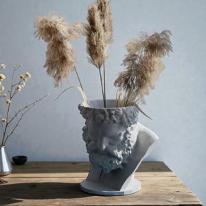 May include: A white matte bust-shaped vase featuring the sculpted head of a bearded figure. The hollow top holds three tall, feathery beige pampas grass stems. The sculpture is positioned on a wooden surface next to a small black bowl.