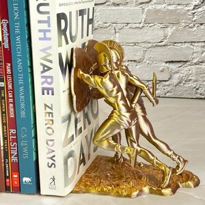 May include: Gold-colored bookends featuring a detailed depiction of two ancient warriors in battle. The bookends are holding up a stack of books, including "Ruth Ware Zero Days" and "The Lion, The Witch and The Wardrobe."
