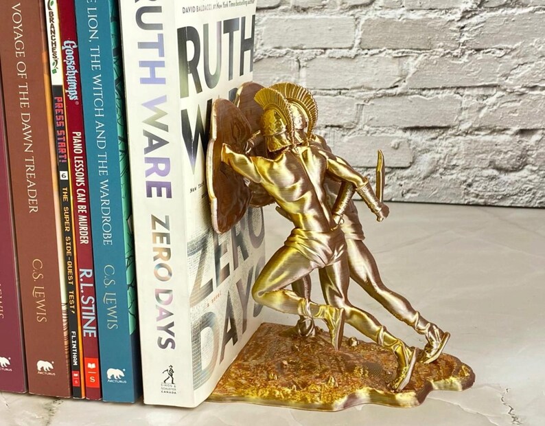 May include: Gold-colored bookends featuring a detailed depiction of two ancient warriors in battle. The bookends are holding up a stack of books, including "Ruth Ware Zero Days" and "The Lion, The Witch and The Wardrobe."
