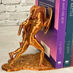 May include: A bronze-colored bookend featuring a sculpted figure of a warrior with a bow and arrow. The bookend is positioned next to a stack of books with titles including "THE AND HIS BOY" and "THE BATTLE".