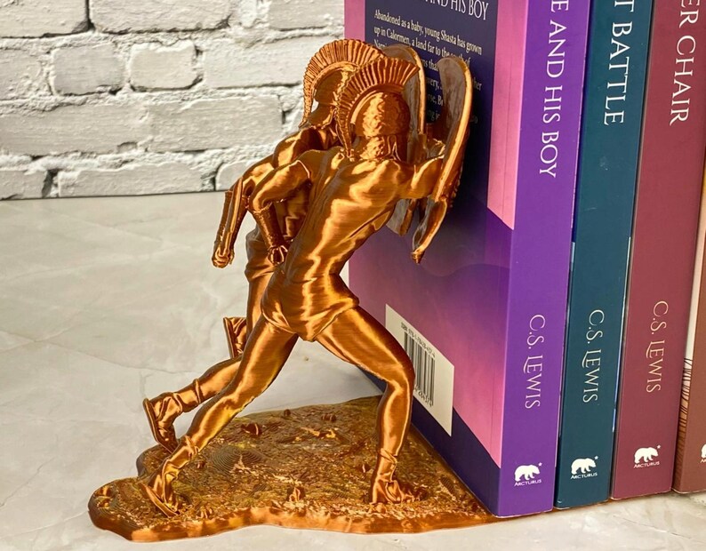 May include: A bronze-colored bookend featuring a sculpted figure of a warrior with a bow and arrow. The bookend is positioned next to a stack of books with titles including "THE AND HIS BOY" and "THE BATTLE".