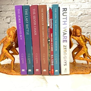 May include: Gold-colored bookends featuring a warrior figure holding a shield and sword, flanking a row of books. The books have titles like "The Horse and His Boy" and "Zero Days". The bookends are approximately 7 inches tall.