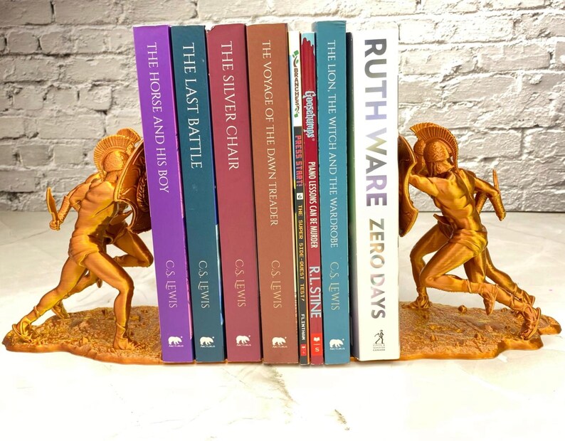 May include: Gold-colored bookends featuring a warrior figure holding a shield and sword, flanking a row of books. The books have titles like "The Horse and His Boy" and "Zero Days". The bookends are approximately 7 inches tall.