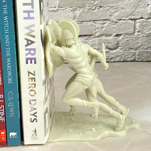 May include: Off-white resin bookend featuring two Spartan warriors in battle. The bookend is holding up a stack of books, including titles like "TH WARE ZERO DAYS" and "THE WITCH AND THE WARDROBE". The bookend adds a decorative touch to any bookshelf.