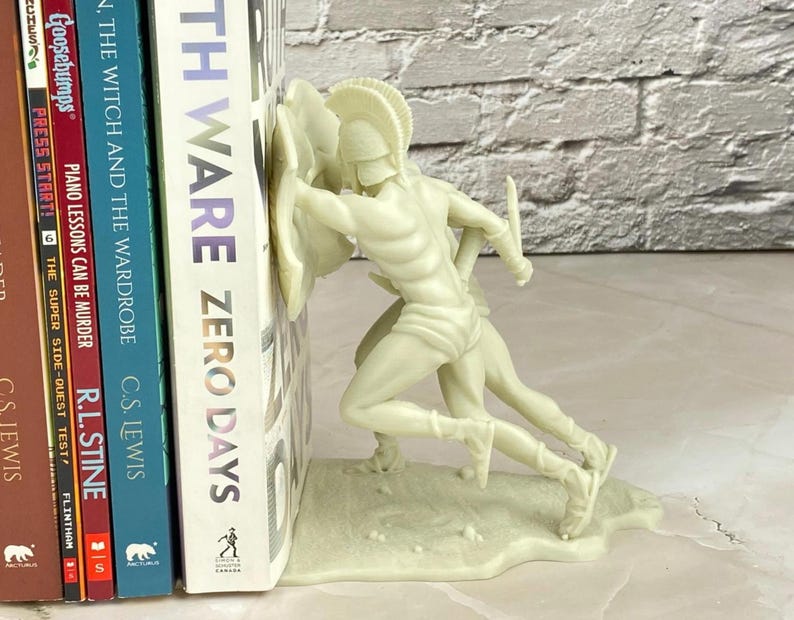 May include: Off-white resin bookend featuring two Spartan warriors in battle. The bookend is holding up a stack of books, including titles like "TH WARE ZERO DAYS" and "THE WITCH AND THE WARDROBE". The bookend adds a decorative touch to any bookshelf.