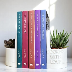 Può includere: Un set di cinque libri colorati di C.S. Lewis, tra cui "Il nipote del mago", "Il cavallo e il suo ragazzo", "La traversata del Viaggiatore dell'Alba", "La sedia d'argento" e "L'ultima battaglia", sono esposti su una libreria bianca con un fermacarte bianco che dice "Booked with Love".