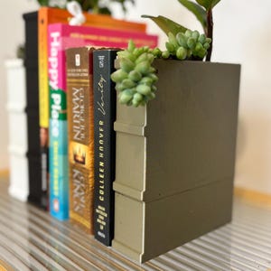Pode incluir: Um vaso decorativo em forma de livro com acabamento dourado metálico, contendo vários livros e pequenas suculentas. Os livros têm lombadas coloridas e títulos como "Happy Place" e "Verity". O vaso adiciona um toque único à decoração da casa.