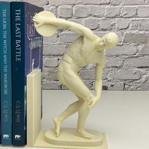May include: Off-white sculpture of a discus thrower, used as a bookend. The figure is in mid-throw, with one arm extended holding the discus. The bookend is placed next to two blue books with the titles "THE LAST BATTLE" and "THE LION, THE WITCH AND THE WARDROBE".