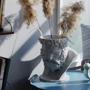 May include: A white sculptural vase shaped like a bearded male head, featuring detailed facial features and hair. The hollow top of the head holds three tall, feathery, light brown dried pampas grass stems, creating a textured and natural display.