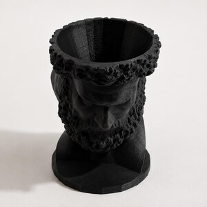 May include: A matte black sculptural planter shaped like a bearded male head. The top of the head is open, creating a cylindrical vessel for plants. The surface features a textured, layered finish consistent with 3D printing, showing detailed facial features.
