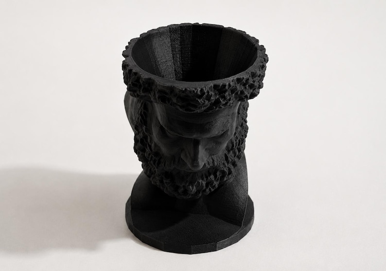 May include: A matte black sculptural planter shaped like a bearded male head. The top of the head is open, creating a cylindrical vessel for plants. The surface features a textured, layered finish consistent with 3D printing, showing detailed facial features.