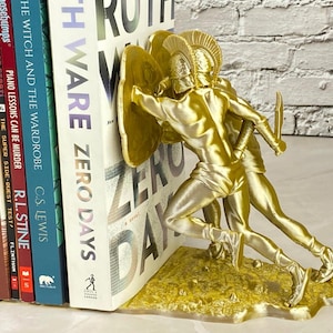 May include: A gold-colored bookend featuring two figures in classical armor, holding shields and swords. The bookend is supporting a stack of books, including "ZERO DAYS" and "THE WITCH AND THE WARDROBE". The bookend is made of a metallic material.