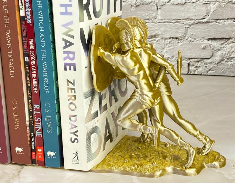 May include: A gold-colored bookend featuring two figures in classical armor, holding shields and swords. The bookend is supporting a stack of books, including "ZERO DAYS" and "THE WITCH AND THE WARDROBE". The bookend is made of a metallic material.