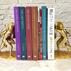 May include: Gold-colored bookends featuring a warrior design, flanking a row of books. The books have titles like "The Horse and His Boy," "The Last Battle," and "Zero Days." The bookends add a decorative touch to any bookshelf.