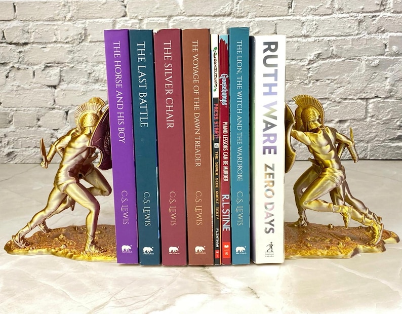 May include: Gold-colored bookends featuring a warrior design, flanking a row of books. The books have titles like "The Horse and His Boy," "The Last Battle," and "Zero Days." The bookends add a decorative touch to any bookshelf.
