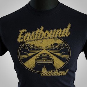 Może przedstawiać: Czarny t-shirt z złotym nadrukiem przedstawiającym samochód jadący drogą z tekstem "Eastbound and down!" nadrukowanym na nim.