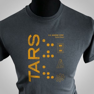 May include: A dark gray t-shirt with a yellow graphic design that says "TARS" and "U.S. MARINE CORP Tactical Robot". The graphic also includes a series of dots and geometric shapes.