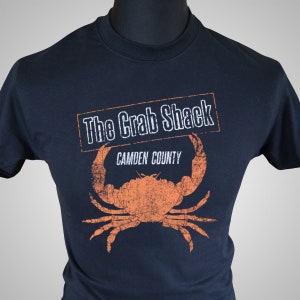 Pode incluir: Uma camiseta azul marinho com um gráfico de caranguejo laranja desbotado e o texto "The Crab Shack Camden County" em branco.