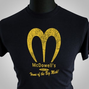 May include: Black t-shirt with a yellow stylized "M" logo and the text "McDowell's" and "Home of the Big Mick!" in yellow. The text "Queens, NY" is in a smaller font below the name.